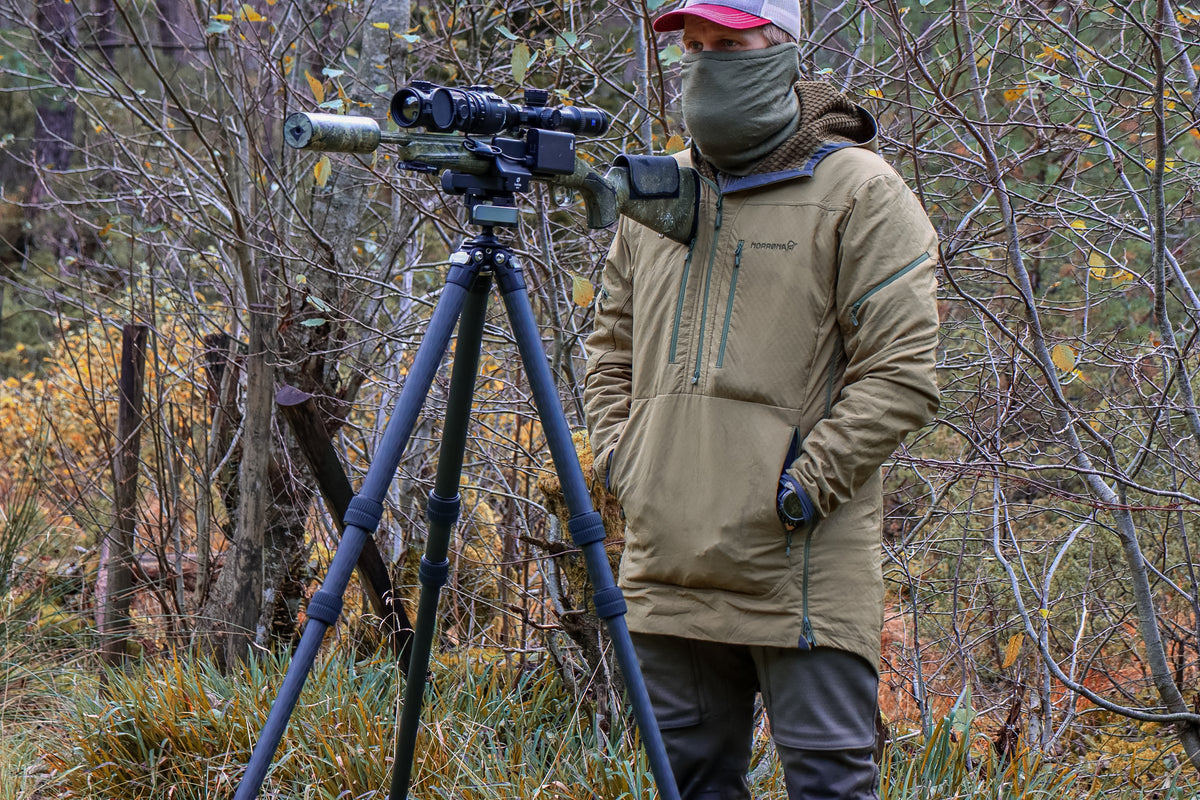 Person standing beside a Leofoto SA-324CL carbon fibre tripod supporting a scoped rifle in woodland.
