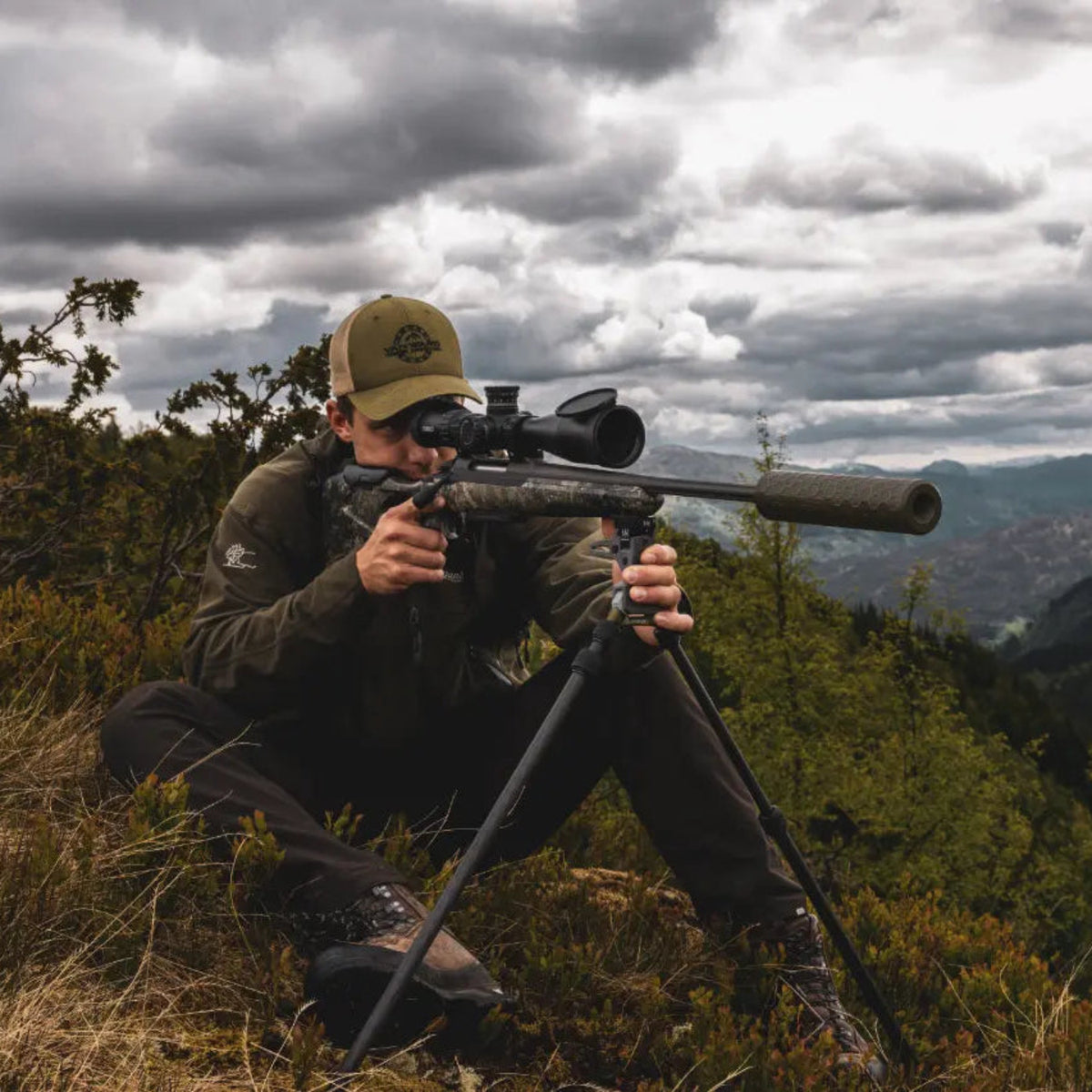 Seated shooter using NorneHunt shooting sticks to support a scoped rifle on a hillside.