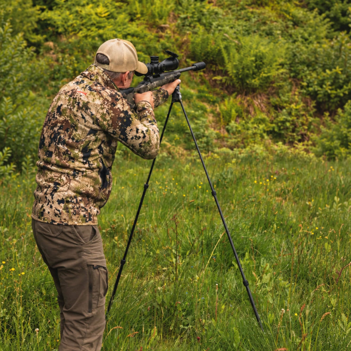 Standing shooter using NorneHunt carbon fibre shooting sticks to support a scoped rifle in a grassy clearing.