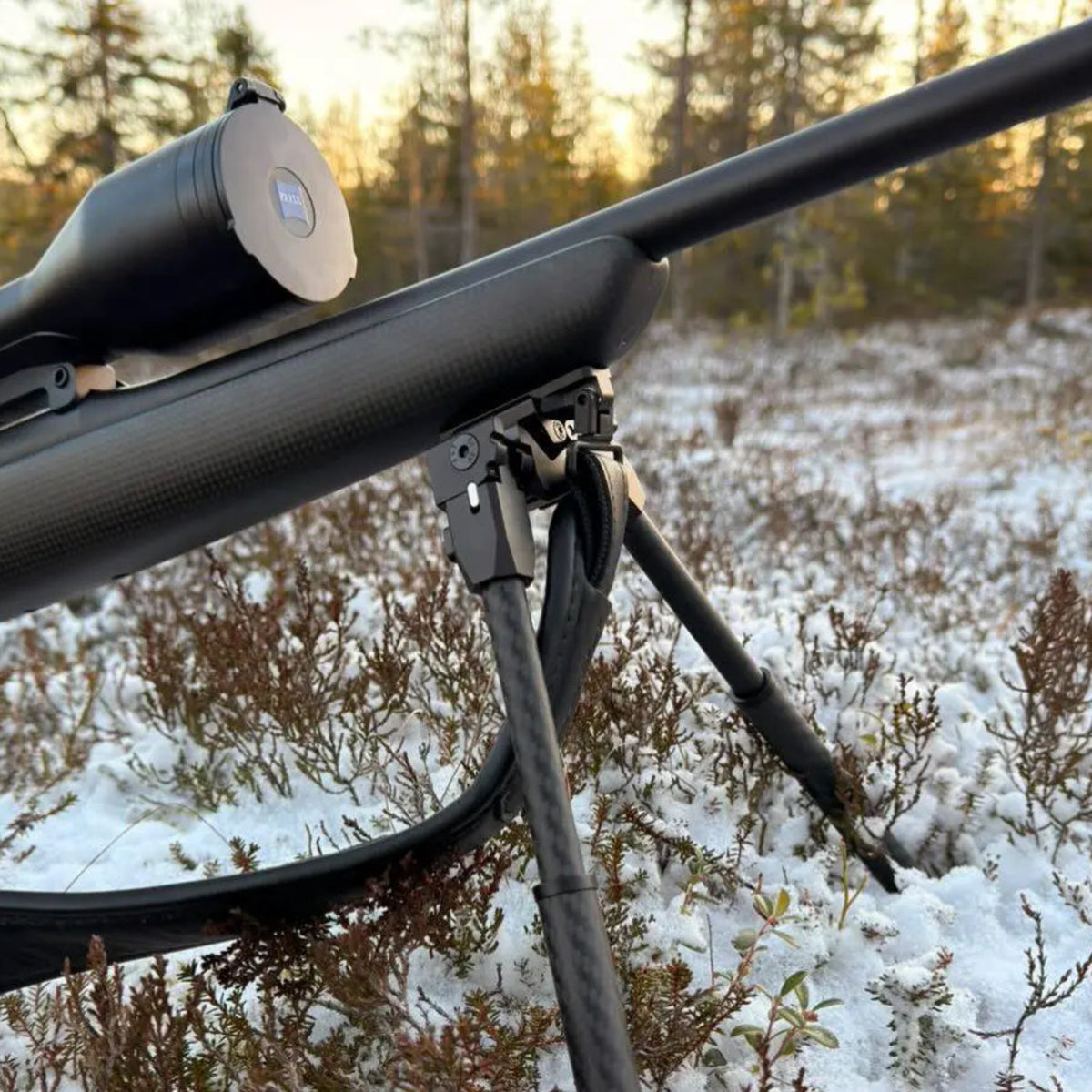 Close-up of a NorneHunt SvartR bipod mounted beneath a rifle with its legs deployed in snow-covered woodland.