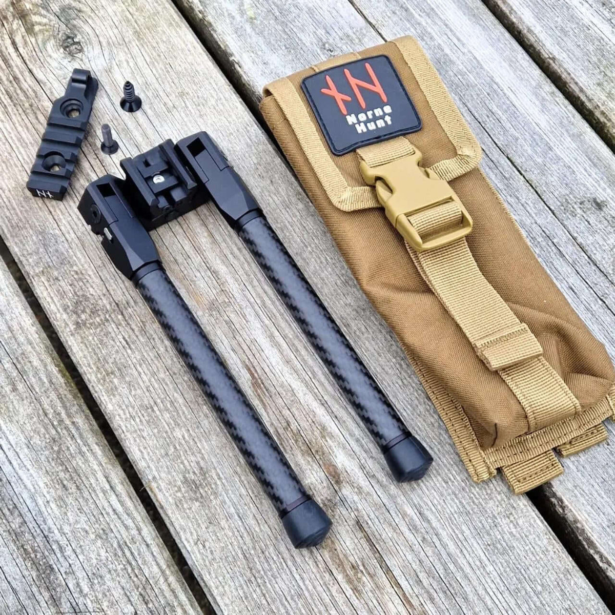 NorneHunt SvartR bipod beside a tan carry pouch, Picatinny rail and two screws on wooden boards.