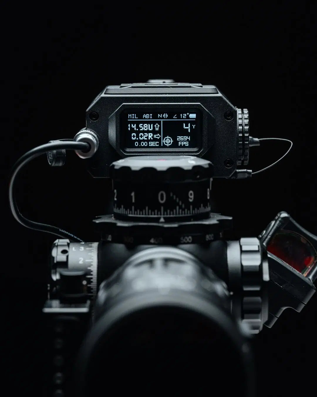 Displayet til TANGO INNOVATIONS FIRE4000 ballistisk avstandsmåler montert på en rifleskinne, med tydelig digital skjerm som viser avstand, vind, ballistiske data og hastighet, omgitt av presise justeringshjul og optikk, alt avbildet mot sort bakgrunn for å fremheve produktets avanserte og funksjonelle design.