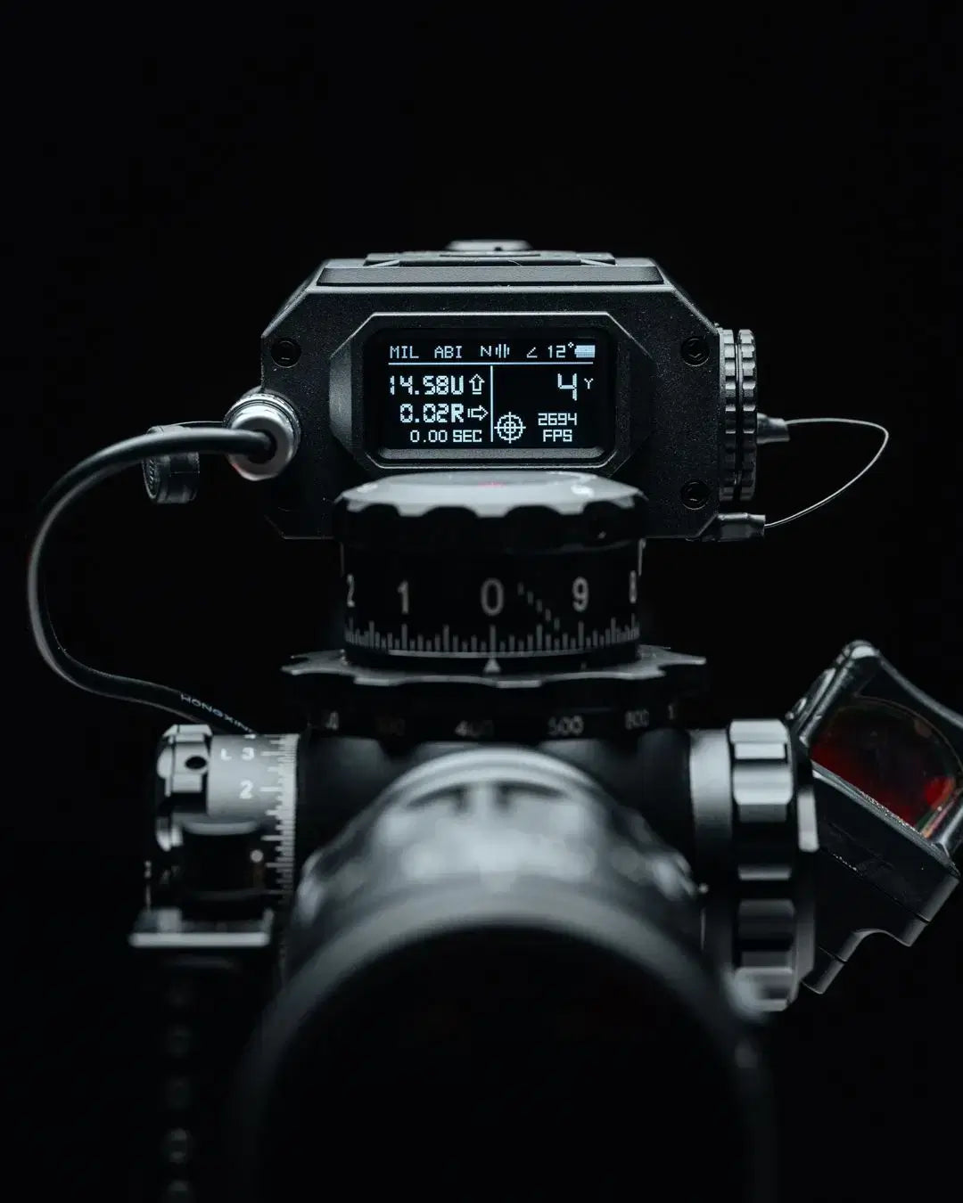 Displayet til TANGO INNOVATIONS FIRE4000 ballistisk avstandsmåler montert på en rifleskinne, med tydelig digital skjerm som viser avstand, vind, ballistiske data og hastighet, omgitt av presise justeringshjul og optikk, alt avbildet mot sort bakgrunn for å fremheve produktets avanserte og funksjonelle design.