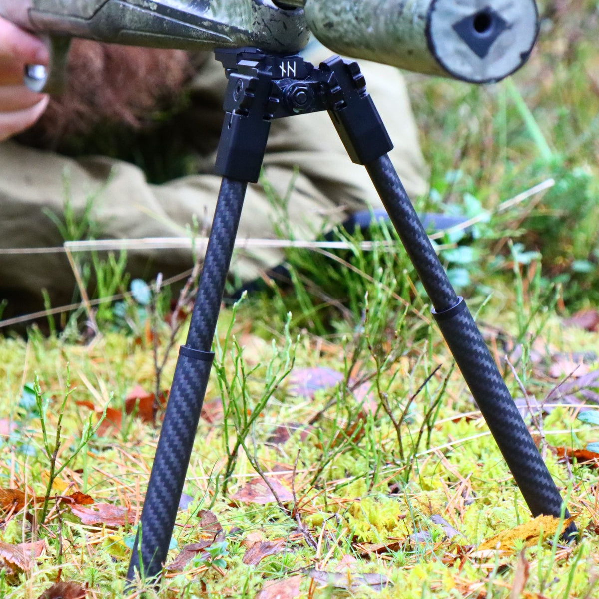 NorneHunt bipod attached beneath a camouflage rifle via a Picatinny rail, with its legs deployed on woodland ground.