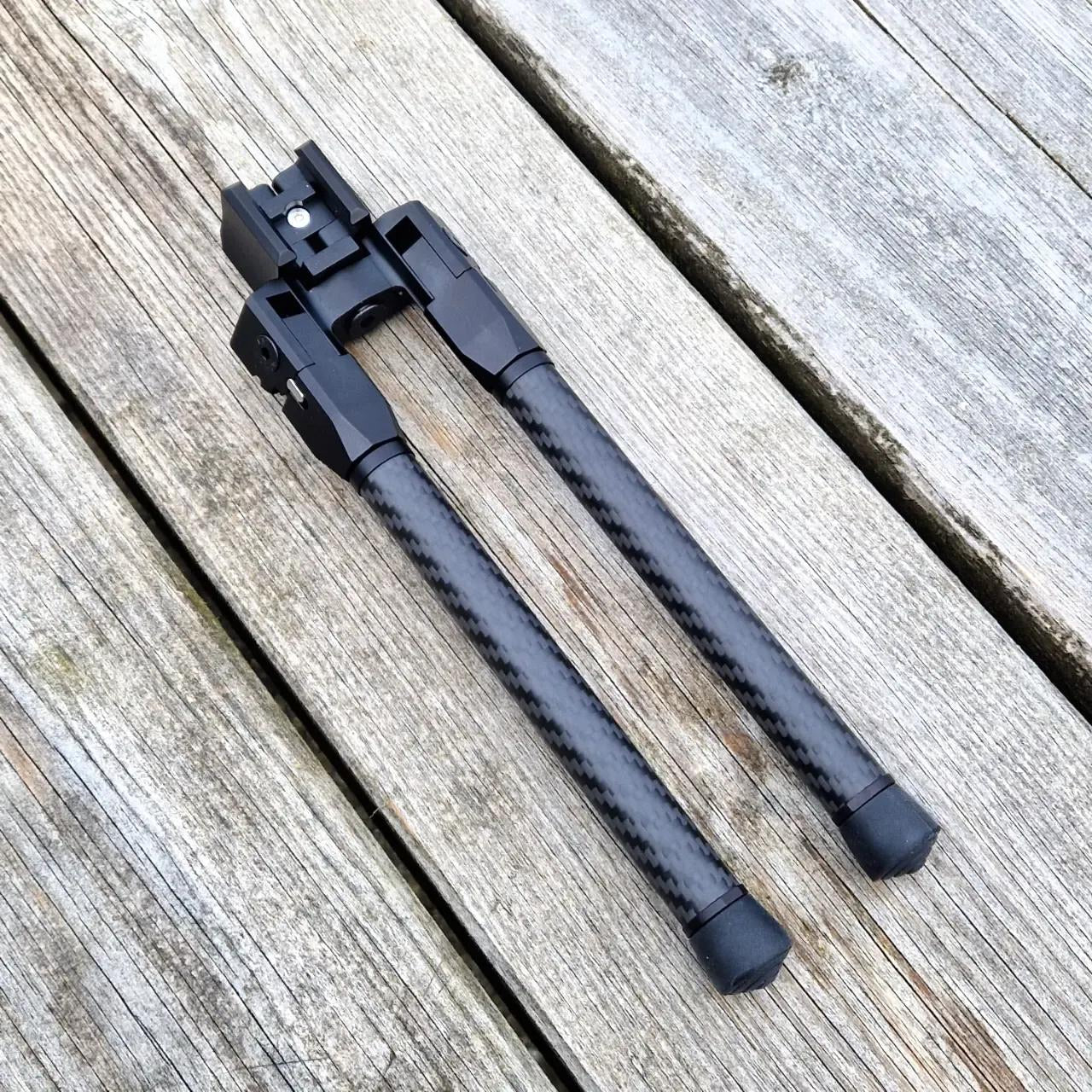 Carbon fiber bipod in a folded position resting on a weathered wooden surface, showing its compact design and sturdy build.