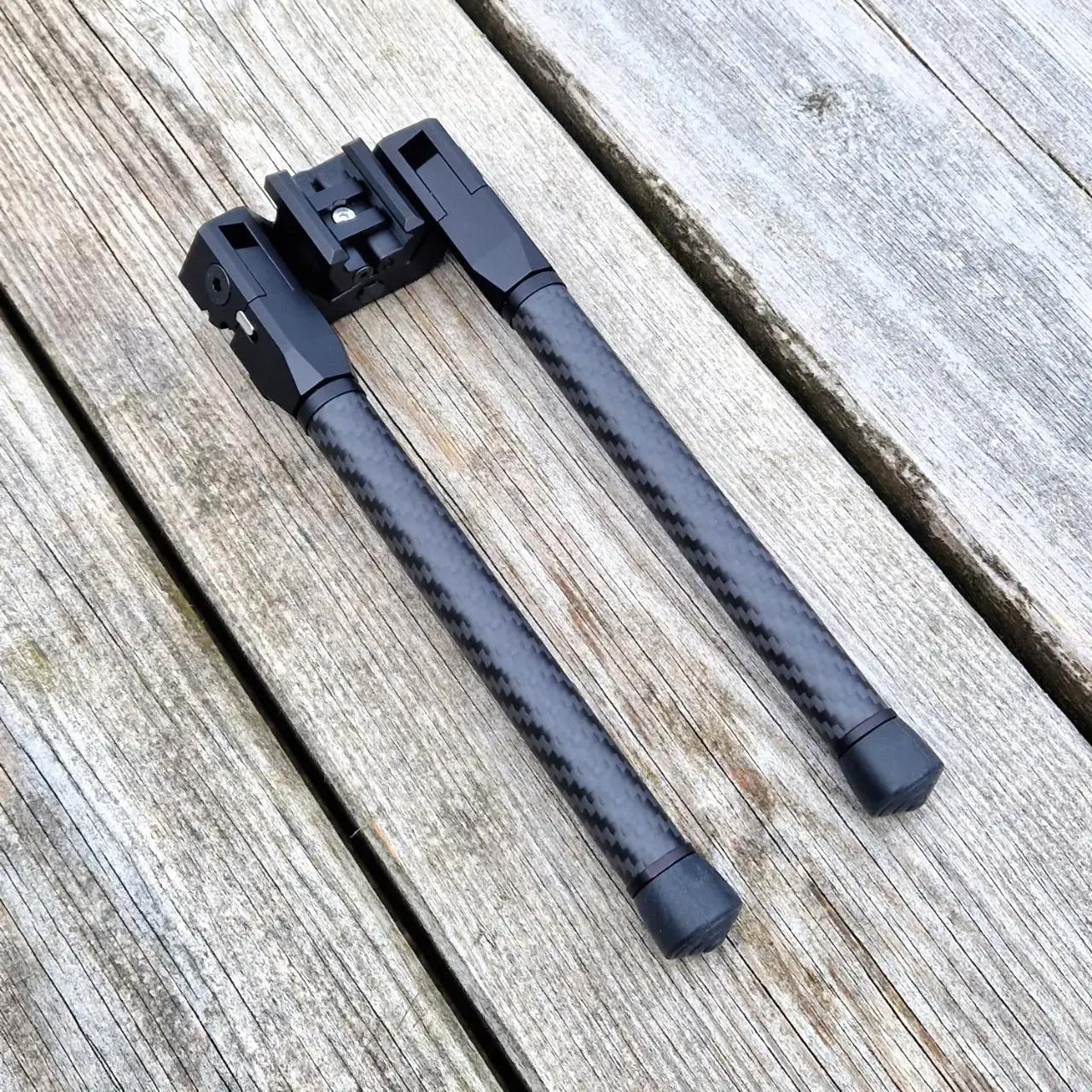 Image displays a close-up of the same bipod with carbon fiber legs, viewed from above and placed on a wooden background.